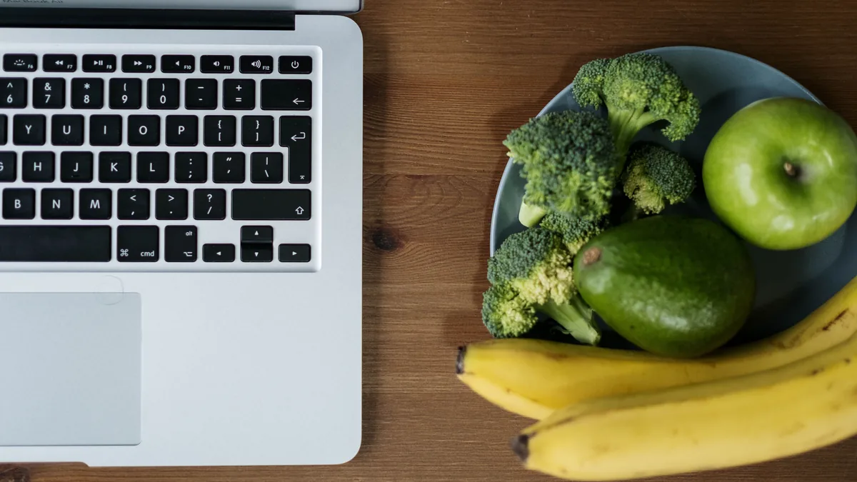Un portatile aperto su un tavolo di legno, accanto a un piatto con mela, avocado, banane e broccoli