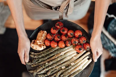 Due mani reggono una teglia scura con pomodorini al forno e asparagi